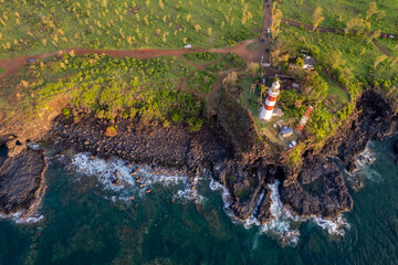 Aerial view of a picturesque lighthouse on a rocky cliff with lush greenery, overlooking the ocean.