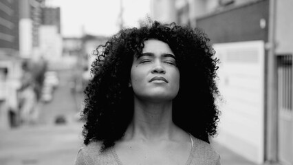 One hopeful young black woman with curly hair standing in street looking up at sky with HOPE and...