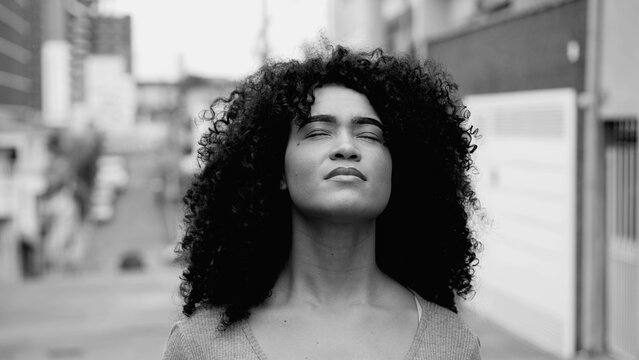 One hopeful young black woman with curly hair standing in street looking up at sky with HOPE and FAITH in black and white. African American person feeling the presence of GOD