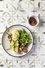 French rolled omelette stuffed with cream cheese with avocado, aragula and mushrooms for breakfast on white plate printed tile background, top view