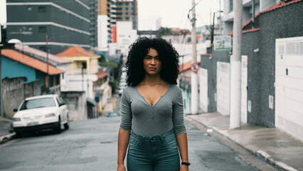 One confident young black woman with curly hair standing outside in urban setting looking at camera...