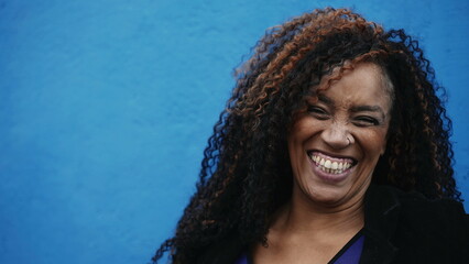 One happy black woman in 40s smiling and laughing at camera on blue backdrop in city street....