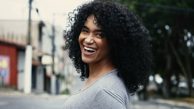 One happy latin black young woman turning head to camera walking in city street laughing and smiling in urban environment, curly haired African American adult girl in 20s