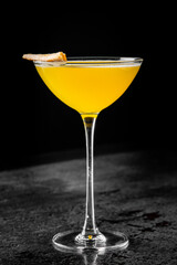 A vibrant yellow cocktail in a martini glass, garnished with an orange slice, against a dark textured background