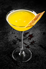 A vibrant yellow cocktail in a martini glass, garnished with an orange slice, against a dark textured background