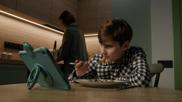 As the boy enjoys his meal and screen time, mom cooks in the kitchen, showcasing modern family dynamics during mealtime