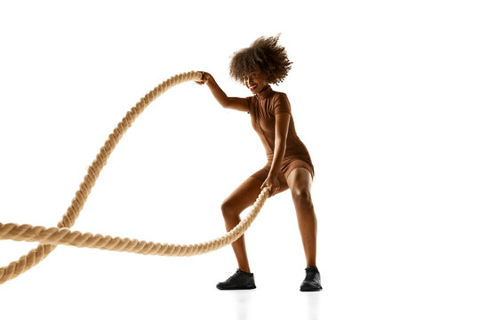 African-American woman in brown workout outfit exercising with heavy battle ropes against white studio background. Concept of sport, mourning routine, active and healthy lifestyle, energy, action.