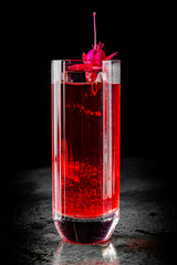 A vibrant red drink in a clear glass, adorned with a bright pink flower, set against a dark textured background