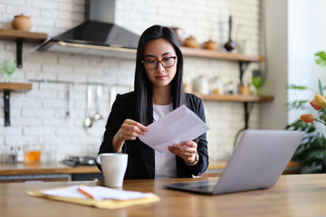 An Asian businesswoman works from home, focused on paperwork and learning, utilizing her laptop and enjoying a cup of coffee.