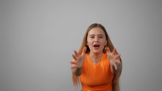 Portrait of young stressed shouting girl. Amazed excited young woman in orange t-shirt screaming raising hands opening mouth closing eyes in studio indoors. Joy, surprise, amazement, panic concept.