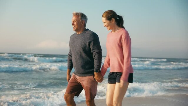 Camera tracks loving retired senior couple on vacation walking along beach shoreline holding hands at sunrise - shot in slow motion
