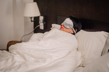 A person is resting in bed with an eye mask on, with a bedside lamp and outlet visible in the...