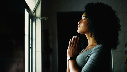 One Spiritual young black woman standing by window at home in prayer with eyes closed. African...