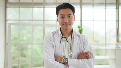 Young asian male doctor in uniform and stethoscope standing with arm crossed smiling to camera. Medical professional with arms crossed with pride for career. Medical and health care concept