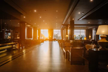Fotobehang Hotel chique An interior view of a luxurious hotel lounge with modern furniture, large windows, and the sun setting in the background casting a warm glow.  © Raivo
