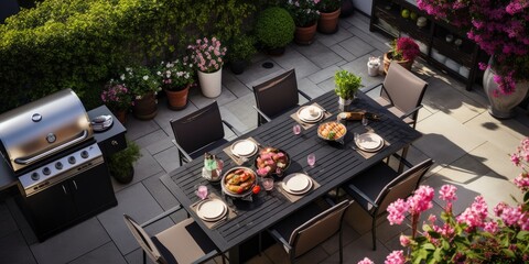 Stylish outdoor kitchen with gas barbecue and dining table set for guests, formal place settings and flowers on paved patio, seen from high angle.