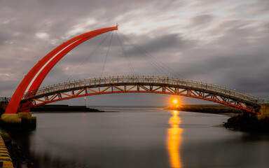 A gloomy sunset in a cloudy evening was sparkled by the light house beneath a red arch bridge