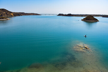 The bright summer sun illuminates the clear water of Lake Kapchagai with a rocky shore, a man on a SUP board sails to a rocky island, an iron bridge is visible in the distance.Kazakhstan