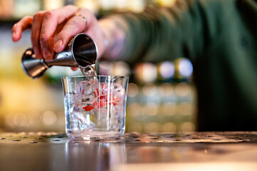 a bartender pouring a clear liquid into a glass with ice and red garnish on a bar counter. The setting appears to be at a bar, with bottles lined up on shelves in the blurred background.