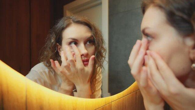 Young happy woman closeup puts on colored contact gray lenses on brown eyes. Adult girl wear lenses stands by mirror looking at reflection smiling face in modern interior bathroom mirror with light 4k