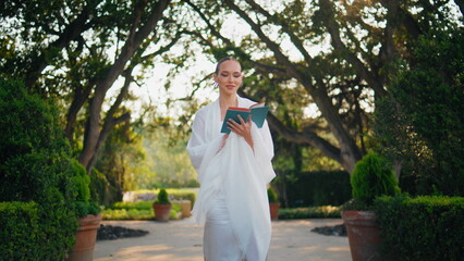 Fototapeta na wymiar Walking woman reading book in sunny garden. Lady smiling enjoying romantic novel