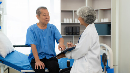 Senior Asian female nurse doctor visits a senior patient sitting on a patient's bed, explaining,...