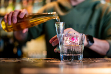 A person pours golden liquid from a bottle into a shot glass on a bar counter. Another glass sits nearby. 