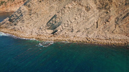 Aerial sunny rocky shoreline washed by turquoise ocean waves super slow motion