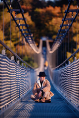 Seated mid-span on a suspension bridge, a young man dressed in warm autumn attire reflects quietly,...