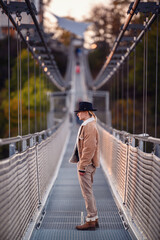 A solitary figure in fashionable autumn attire pauses for a reflective moment on a suspension bridge, with the early evening light accentuating the fall setting