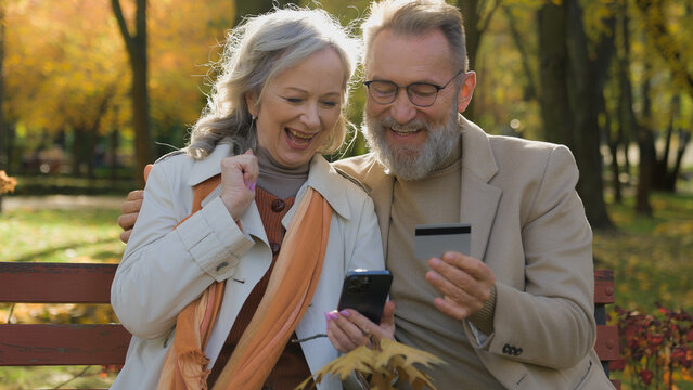 Caucasian couple happy 60s old mature woman man hold credit card mobile phone bank app two grandparents in city park shopping online purchase pay money with smartphone celebrate successful transaction