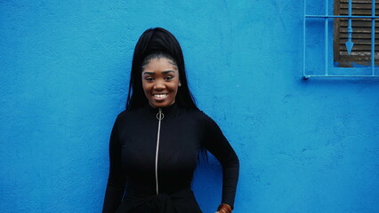 One young black woman standing outside in urban environment. 20s African American adult girl smiling on blue wall backdrop, portrait face
