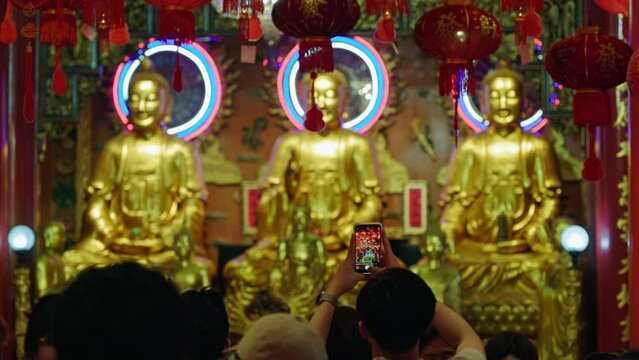 In a richly decorated temple, a visitor captures golden Buddha statues adorned with colorful halos on a smartphone, surrounded by red Chinese lanterns and other worshippers. Bangkok Thailand. 5.01.202