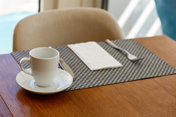 A white cup and saucer sit on a woven placemat on a wooden table. Beside them, a folded white napkin and silver fork are neatly placed. The chair in the background and natural light through the window