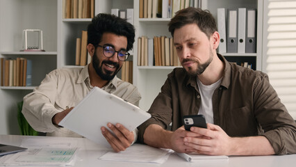 Two businessmen multiracial men co-workers in office Arabian Indian businessman show papers business project on laptop to colleague Caucasian man ignoring addicted mobile phone inefficient teamwork - obrazy, fototapety, plakaty