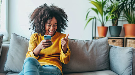 Happy young black woman sitting on sofa at home with phone in her hand. - obrazy, fototapety, plakaty