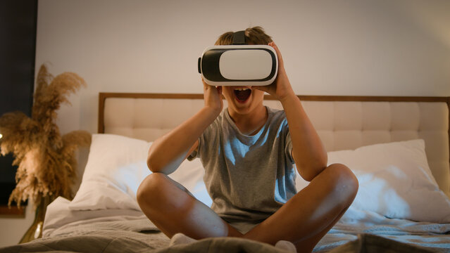 Surprised amazed Caucasian child kid boy playing video game using innovative digital VR headset on bed home play online 3d AR technology in metaverse world wearing wireless virtual reality glasses - Powered by Adobe