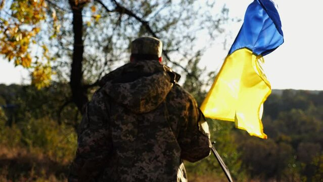 Young male military in uniform walks with waving flag of Ukraine at countryside. Soldier of ukrainian army going with blue-yellow banner outdoor. Concept of victory against russian aggression