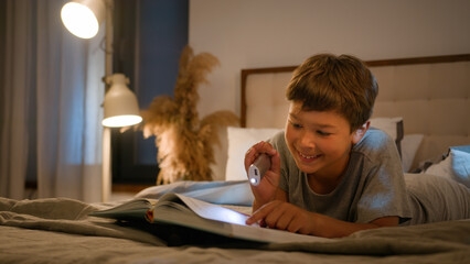 Surprised amazed smiling funny Caucasian child kid boy schoolboy reading interesting book evening...