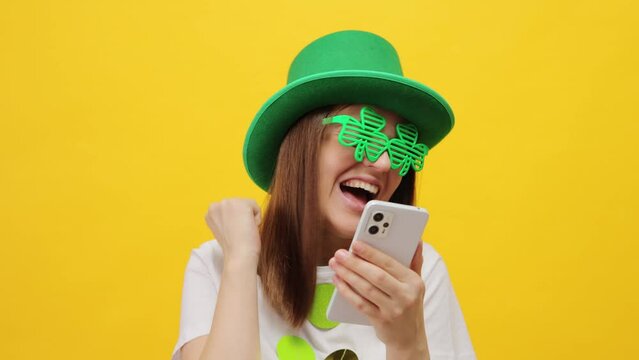 Pretty extremely happy woman wearing leprechaun party green hat and clover glasses isolated over yellow background using mobile phone clenched fist rejoicing good news