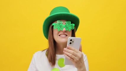 Attractive smiling delighted woman wearing leprechaun party green hat and clover glasses isolated over yellow background using cell phone for online communication has video call waving hand hi gesture