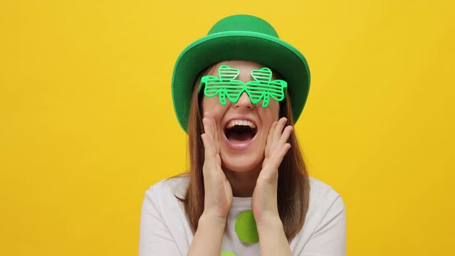 Happy joyful St. Patrick's Day leprechaun model female in green hat and clover  glasses isolated over yellow background keeps hands near mouth exclaiming screaming making announce