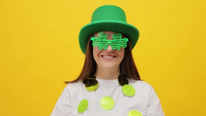 Smiling cheerful St. Patrick's Day leprechaun model female in green hat and clover  glasses celebrating holiday with positive emotions isolated over yellow background