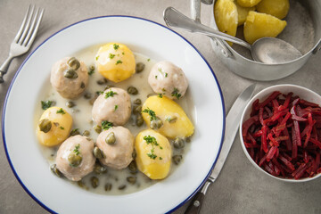 Königsberger Klopse mit Kapern, Salzkartoffeln und Rote Beete Salat auf rustikal Emaille Teller und grauen Stein Hintergrund mit Besteck, Salz und Serviette
