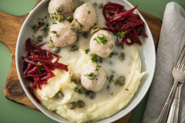 Königsberger Klopse mit Kapern, Kartoffelpüree und Rote Beete Salat in Schale mit  Holzbrett, Silber Besteck und Serviette auf Hintergrund grün