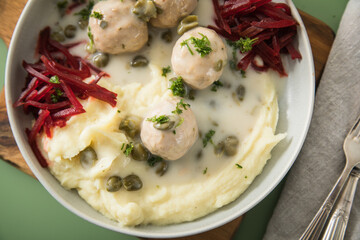 Königsberger Klopse mit Kapern, Kartoffelpüree und Rote Beete Salat in Schale mit  Holzbrett, Silber Besteck und Serviette auf Hintergrund grün