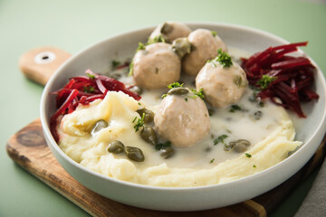 Königsberger Klopse mit Kapern, Kartoffelpüree und Rote Beete Salat in Schale mit  Holzbrett, Silber Besteck und Serviette auf Hintergrund grün