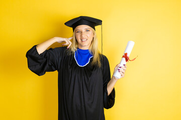 Beautiful blonde young woman wearing graduation cap and ceremony robe covering ears with fingers...