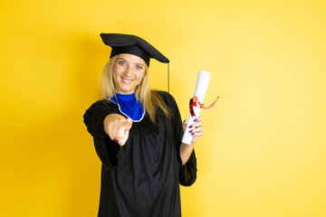 Beautiful blonde young woman wearing graduation cap and ceremony robe laughing at you, pointing...