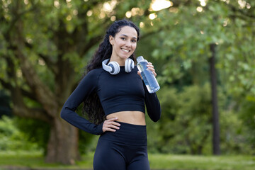 Joyful fitness enthusiast takes a rest in a green park, holding a water bottle with headphones...